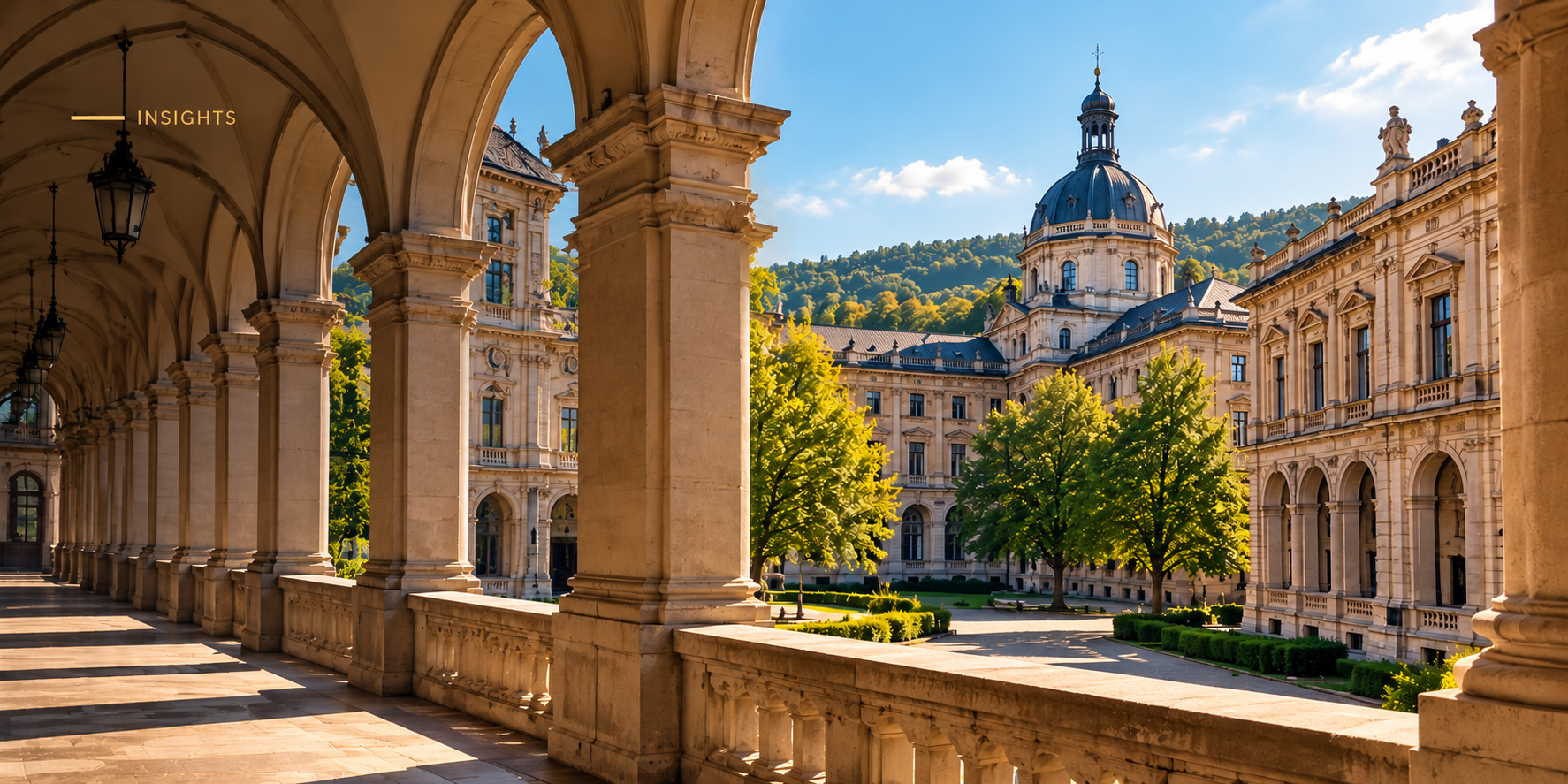 Sunlit stone arcade with columns and a balustrade, looking out onto a baroque university building with a domed tower, green trees and wooded hills under a blue sky.