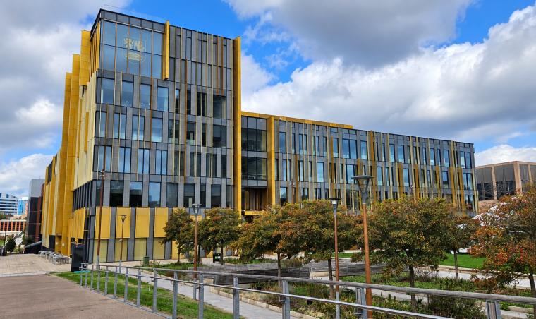 Modern university building with a glass-and-gold facade and an emblem above the entrance, under a partly cloudy sky.